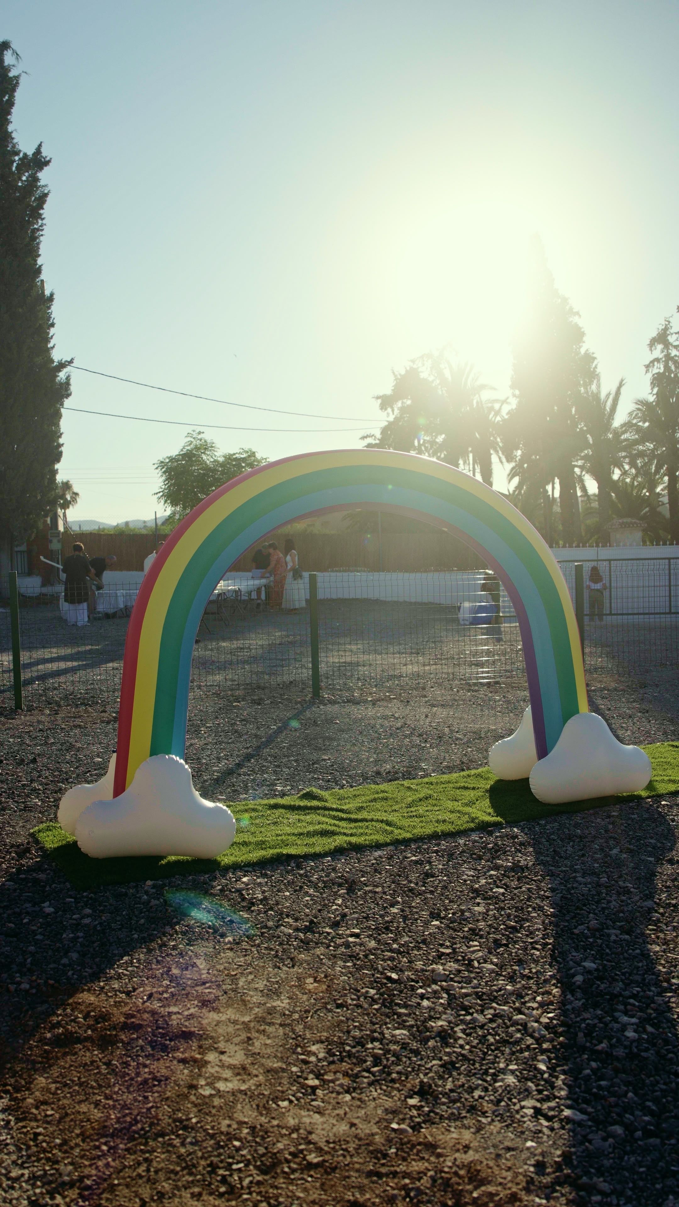 Un arco hinchable con los colores del arcoíris y bases con forma de nube, montado sobre una franja de césped artificial; al fondo, árboles, una valla blanca y personas a contraluz.
