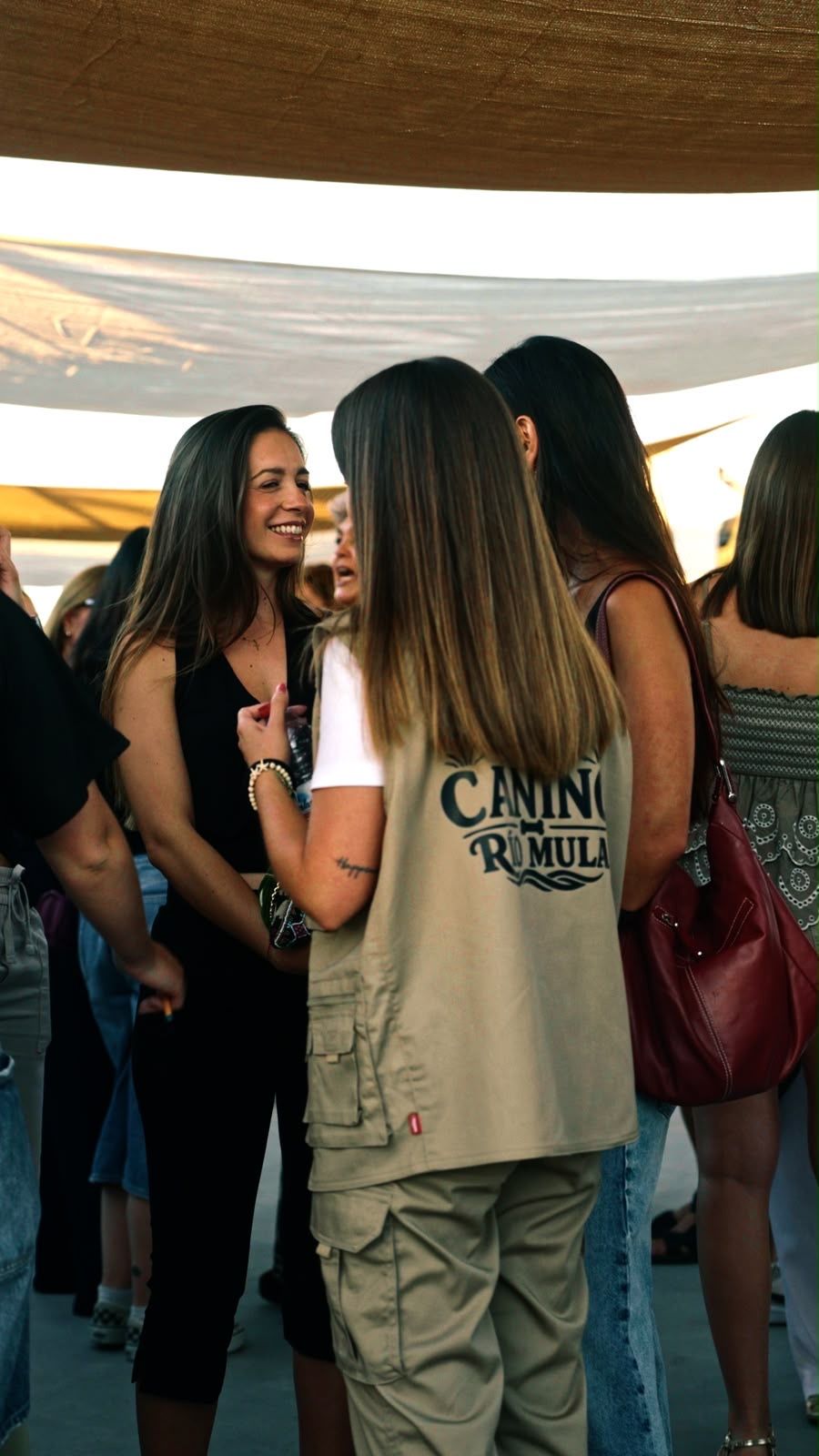 La misma escena bajo la carpa en un encuadre más abierto: dos personas conversan sonriendo y se ve el resto del grupo alrededor.