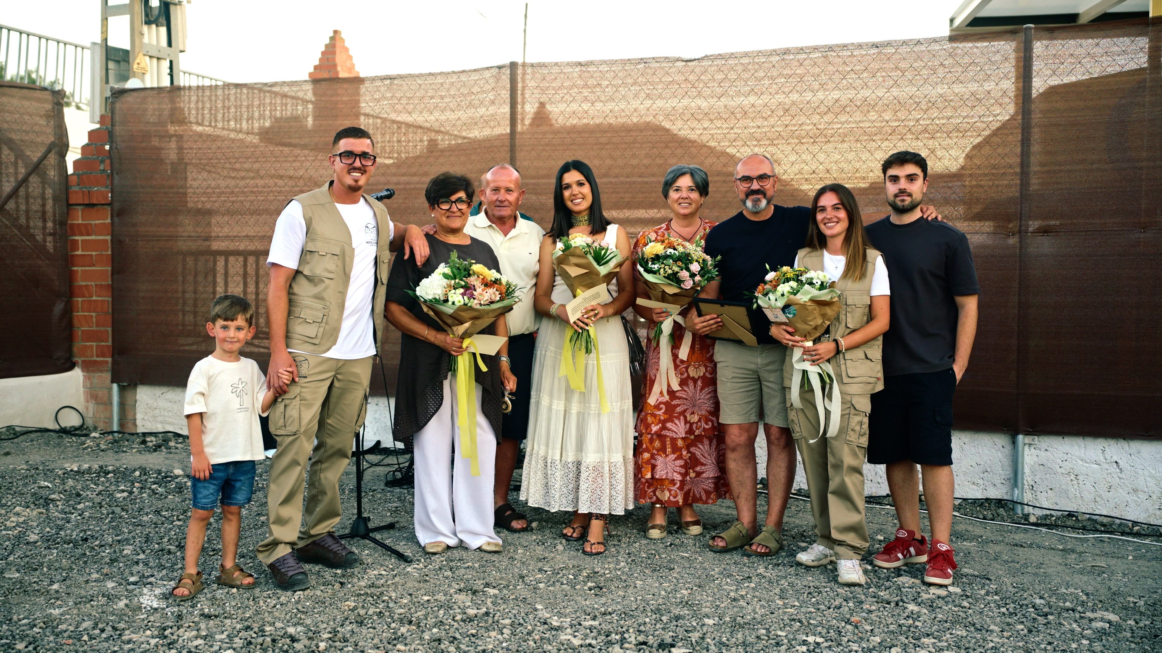 Grupo de nueve personas posando en fila ante una valla de ocultación marrón con pilares de ladrillo; cuatro sostienen ramos de flores. Suelo de grava.