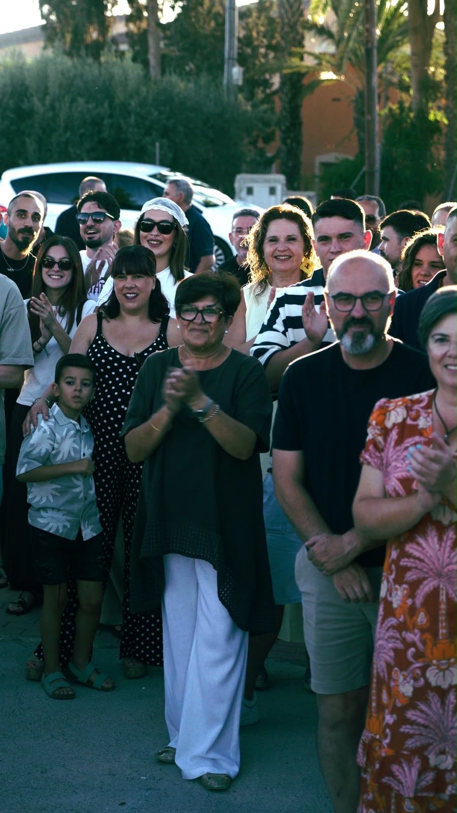 Un grupo de personas aplaude al aire libre al atardecer, con coches aparcados y árboles al fondo.