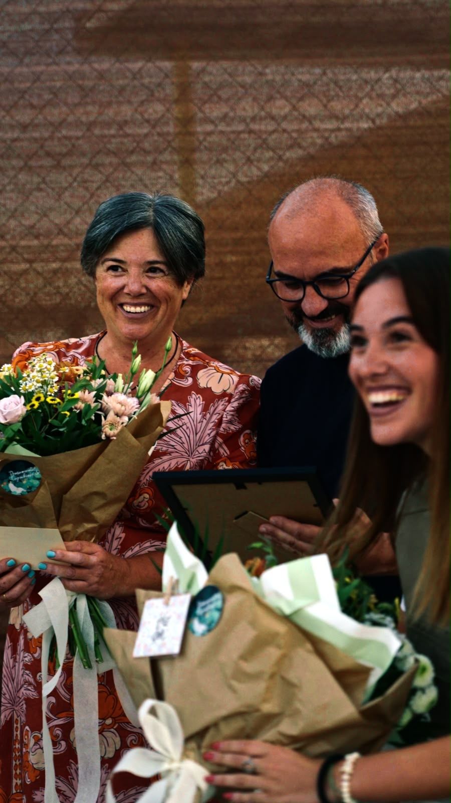 Tres personas sonríen tras una entrega: dos sostienen ramos de flores envueltos en papel y una tercera mira una placa enmarcada.
