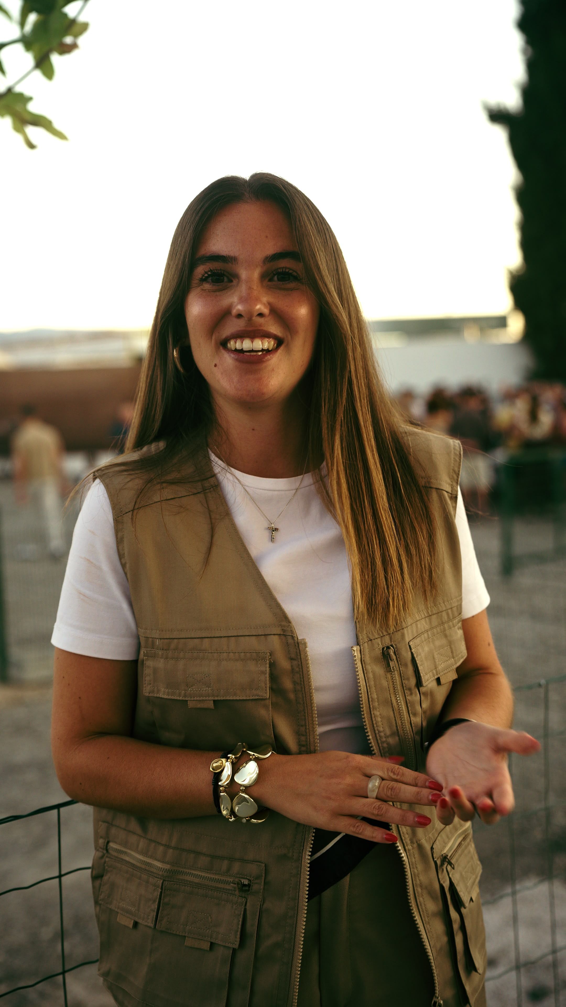 Una persona con camiseta blanca y chaleco beis sonríe a cámara con una mano abierta hacia delante; detrás, a contraluz, una valla metálica y gente desenfocada.