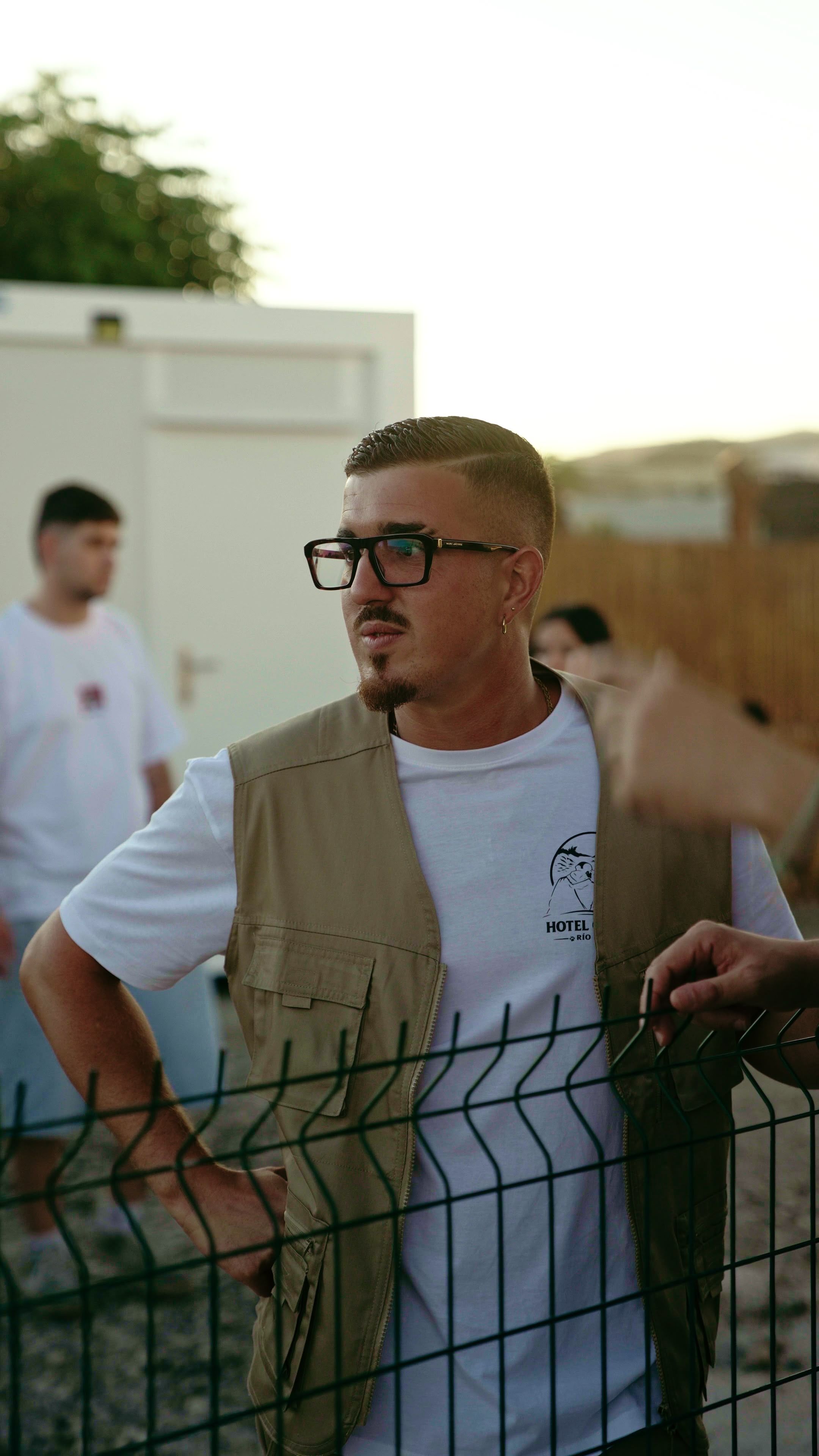 Una persona con gafas, camiseta blanca serigrafiada y chaleco beis de bolsillos, apoyada en una valla metálica verde al atardecer; detrás, otras personas desenfocadas.