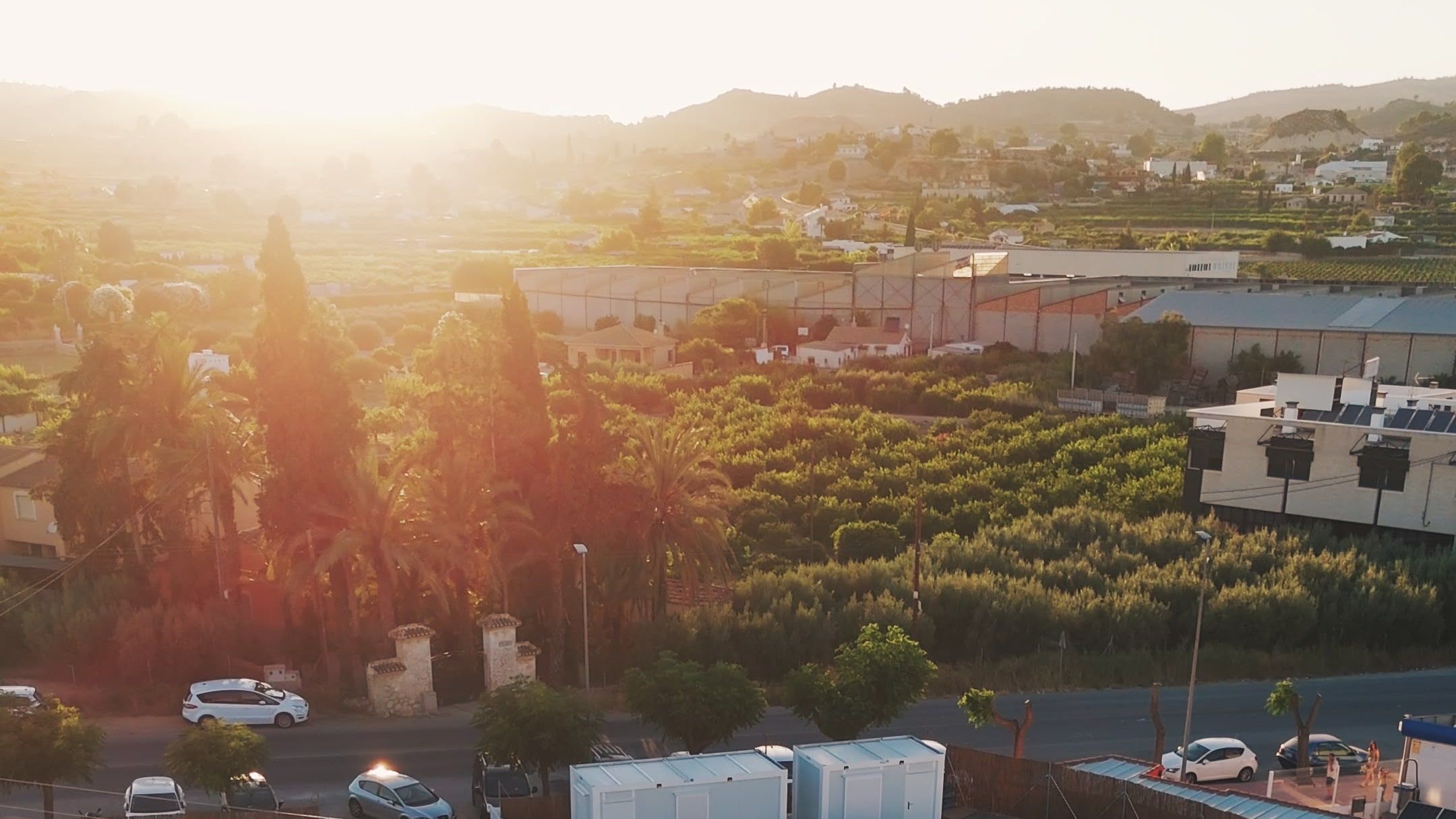 Vista aérea al atardecer del recinto vallado, con módulos y velas de sombra, rodeado de campos de cultivo y montañas.